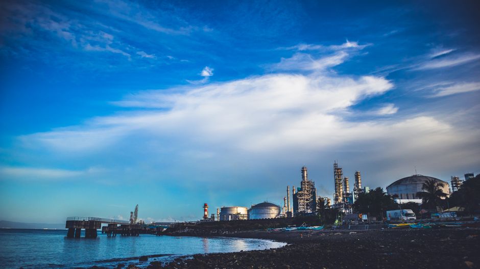 Industrial refinery buildings along a coastline under a dramatic cloudy sky, illustrating energy infrastructure exposed to geopolitical risk.