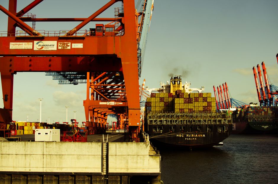 A container ship is handled by cranes at an industrial port as global trade faces longer routes and higher shipping costs.