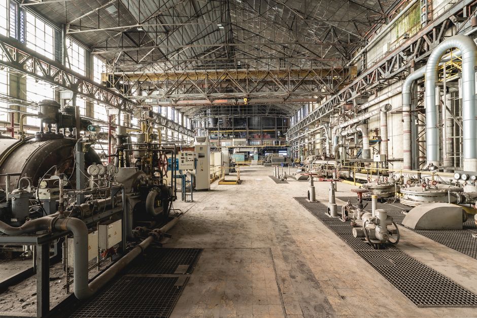 Spacious industrial facility interior featuring machinery and a network of large pipes running along the walls and ceiling.
