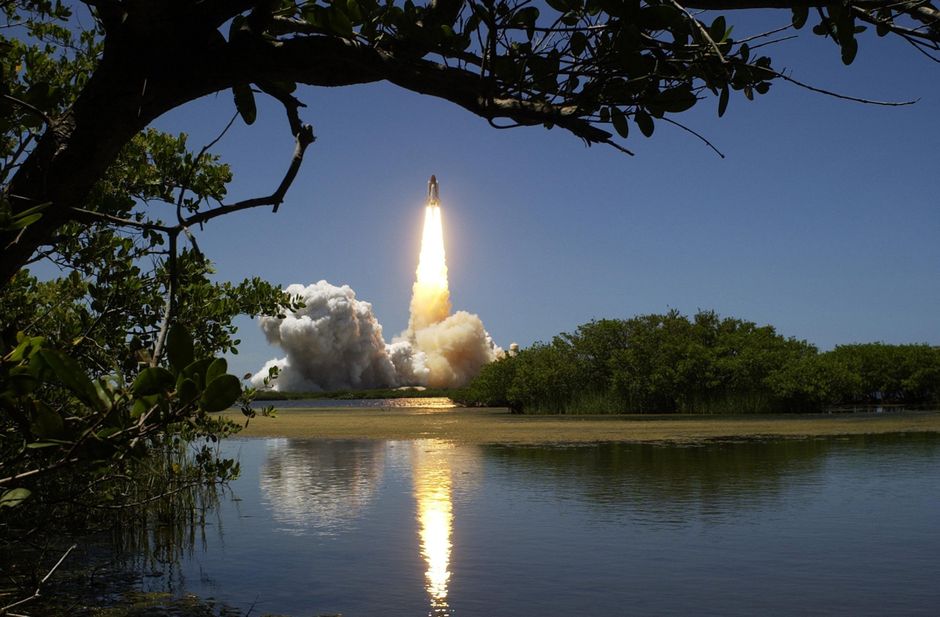 A rocket launching with smoke and flames, reflected in calm water and framed by trees