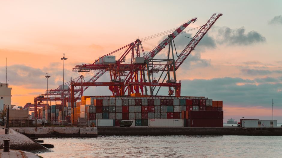Container cranes at a bustling industrial shipping port during sunset, representing the Strait of Hormuz energy chokepoint.