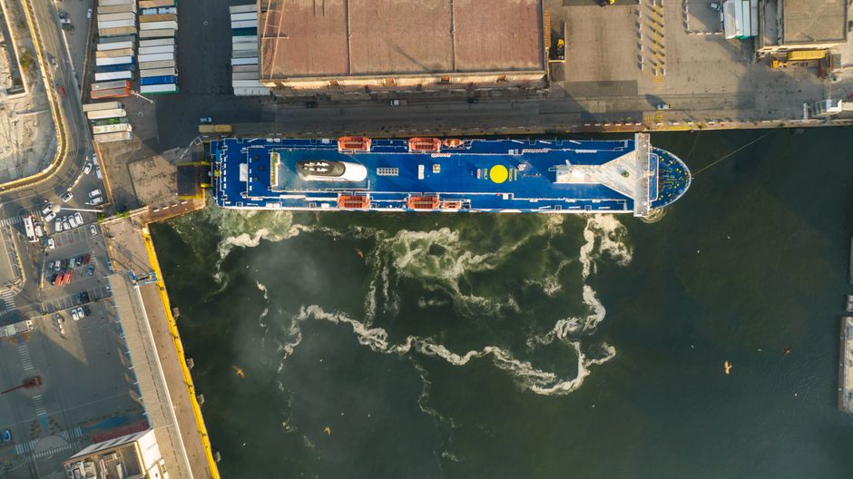 Aerial view of a large cargo ship docked at an industrial port with visible harbor infrastructure, representing maritime energy trade through strategic shipping chokepoints.