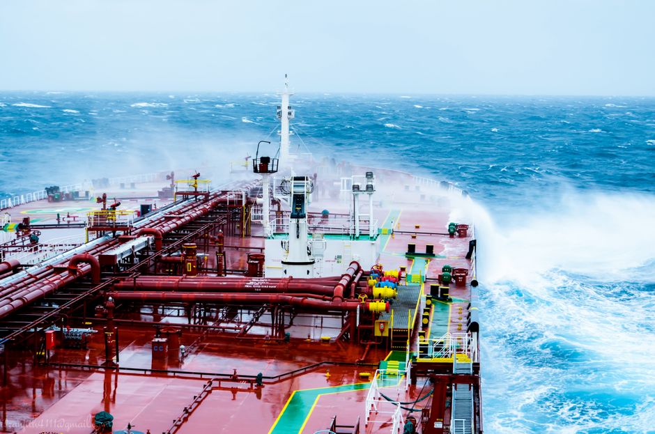 A cargo ship navigating through rough ocean waves on a bright day, illustrating the maritime oil shipping routes now disrupted by dual chokepoint closures in the Strait of Hormuz and Bab al-Mandeb.