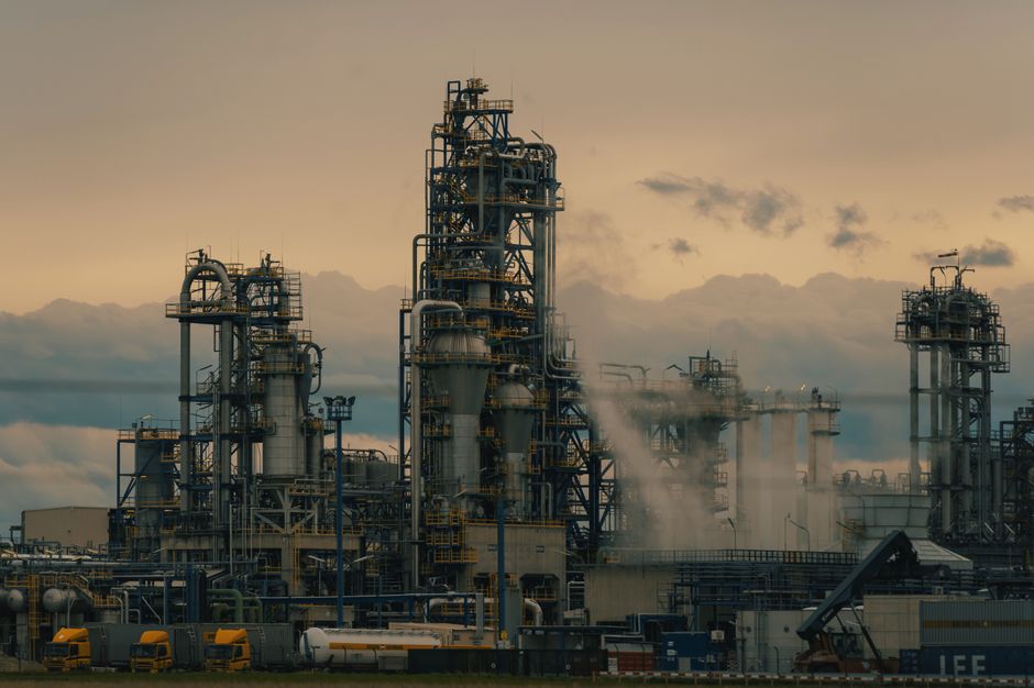 Oil refinery towers silhouetted against a cloudy dusk sky, illustrating the energy supply chain at the center of the US-Iran crisis