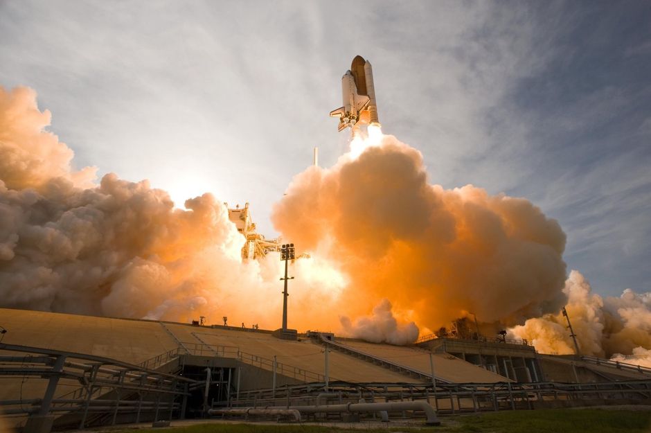 A space shuttle launching upward surrounded by smoke and fire against a clear sky.