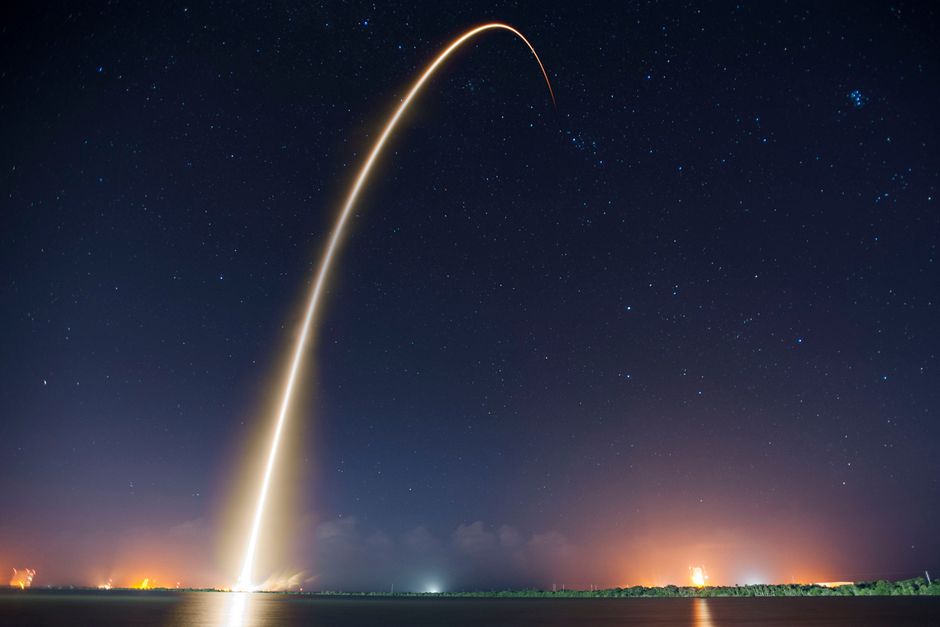 A long-exposure photograph of a rocket launching at night, leaving a bright trail of light against a starry sky.