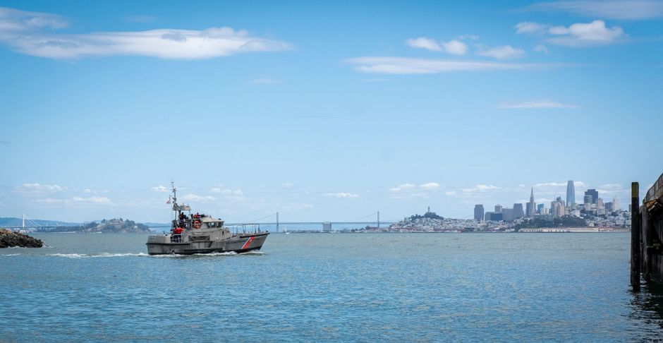 Naval vessel patrolling coastal waters with a city skyline and bridges in the background.
