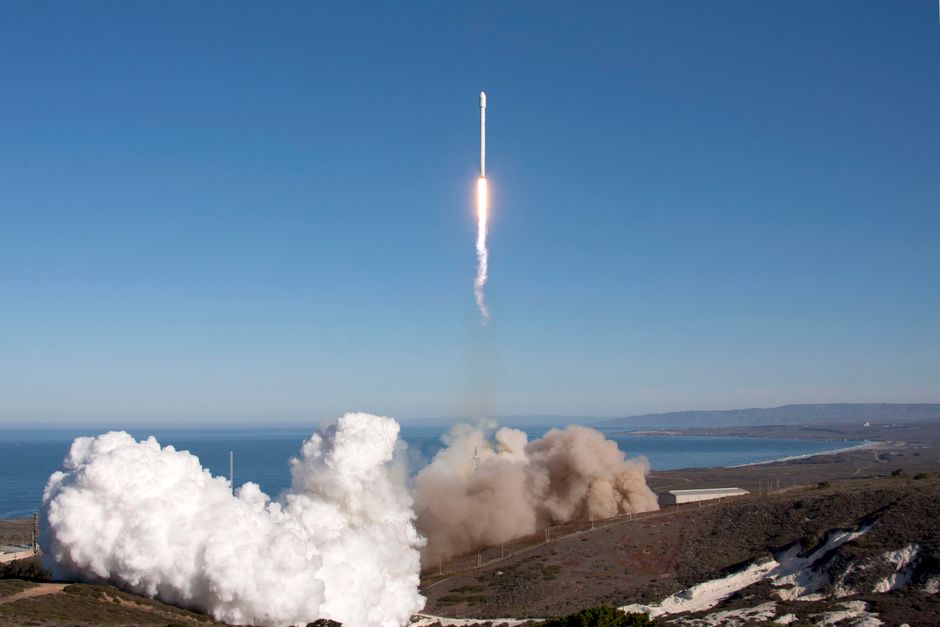 A rocket launches over the ocean, leaving behind smoke and fire trails against clear skies.