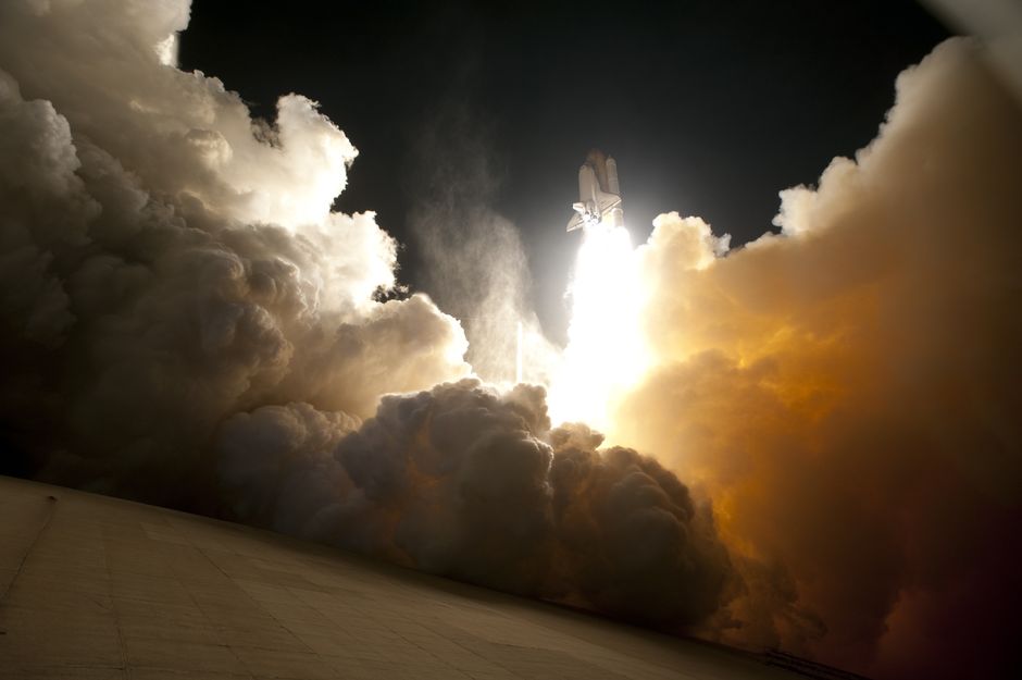 A rocket launches into a night sky, illuminating billowing exhaust clouds below.