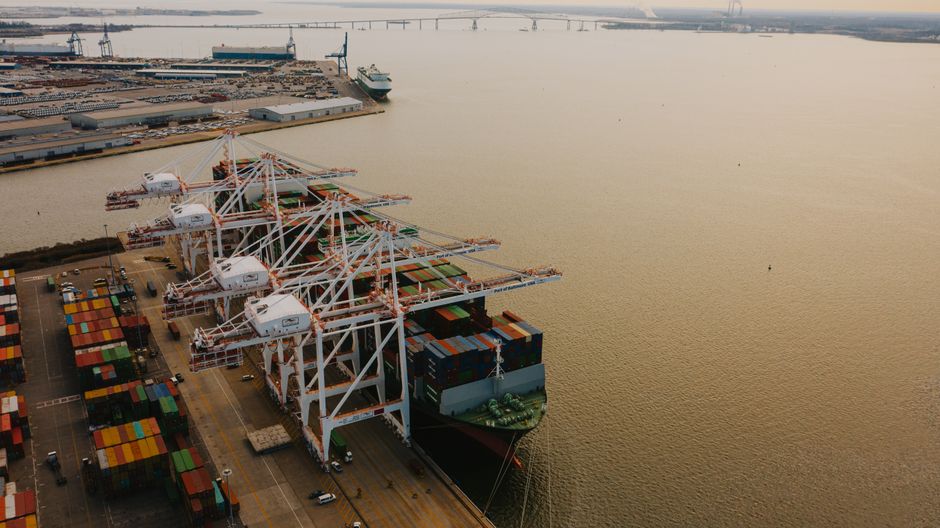 Aerial drone view of a large cargo ship being loaded with shipping containers by cranes at a port.