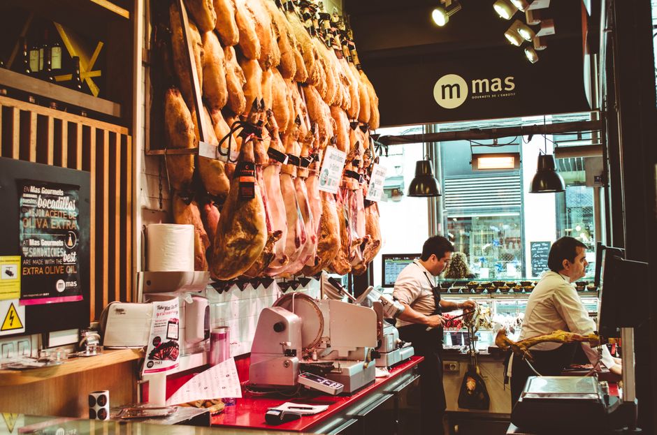 A busy deli shop interior with hanging cured meats and staff serving customers at a counter