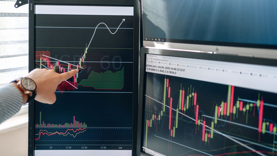 Person pointing at stock market graphs on dual monitors in a modern trading workspace
