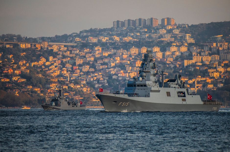 Naval warships patrol coastal waters near a inhabited shoreline, underscoring militarized maritime chokepoints.