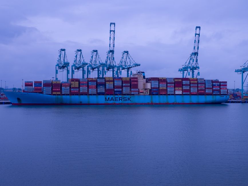 A large Maersk cargo ship loaded with containers docked at an industrial port.