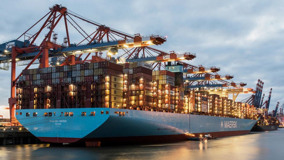 View of a large cargo ship being loaded at harbor during twilight, illustrating global trade and energy shipping vulnerability at maritime chokepoints