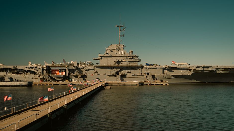 Aircraft carrier docked at harbor with walkway and flags, representing U.S. naval military presence amid escalating Iran conflict
