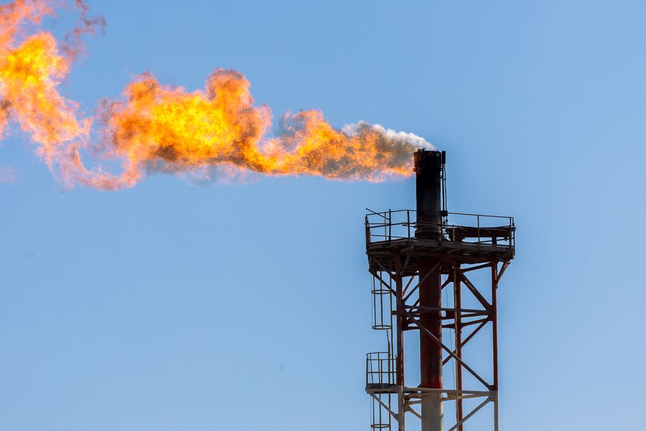 Industrial gas flare tower with bright orange flame against a clear sky.